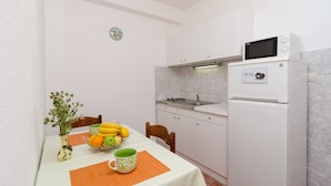 Une cuisine compacte avec des meubles blancs, un four à micro-ondes et un évier. On y trouve une petite table à manger avec un saladier de fruits et une tasse.