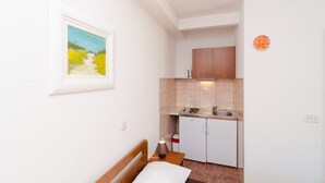 Une petite pièce, de taille modeste, comprenant un lit, une table de chevet, un petit coin cuisine et un tableau accroché au mur.