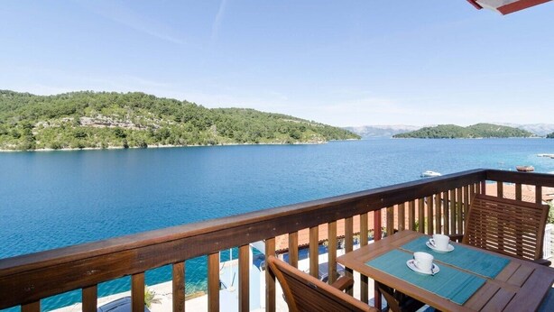 Una terraza de madera con una mesa y sillas que da a un lago con barcos, rodeada de colinas.