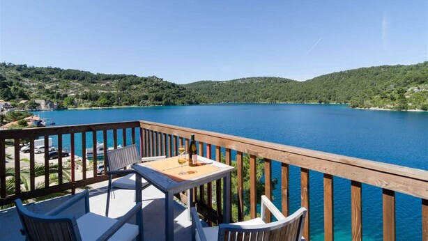 A wooden deck with a table and chairs overlooking a lake with a forested hillside in the background. - Corso turquoise view, 1BR APT with balcony and sea view (Mljet)