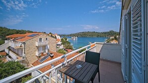 Un balcon avec une table et des chaises donnant sur une marina où sont amarrés des bateaux, un ciel bleu dégagé et, au loin, un paysage vallonné.