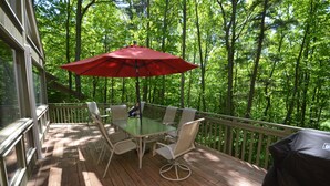 A wooden deck with a glass table and chairs, a red umbrella, and a view of a forest.