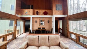 A spacious living area with a large leather sofa, wooden ceiling, and large windows offering views of the surrounding forest.