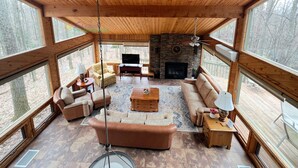 A spacious living room with a stone fireplace, wooden ceiling, and large windows offering a view of the forest.
