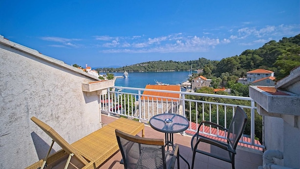Un balcon avec des fauteuils en osier et une petite table, donnant sur une étendue d’eau où l’on aperçoit des bateaux et des bâtiments au loin.
