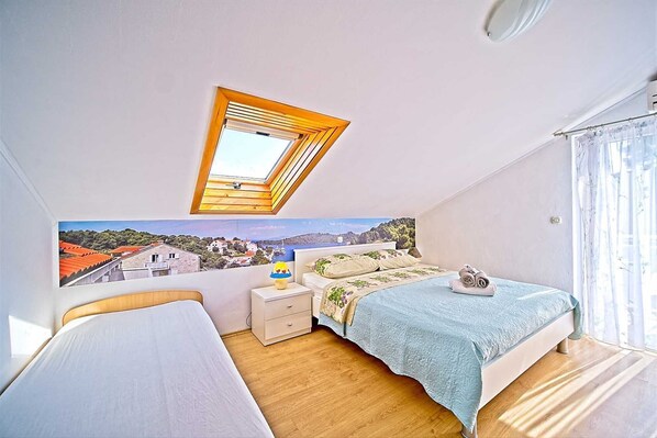 A bedroom with a skylight, a bed with a blue bedspread, a nightstand, and a view of a coastal town.