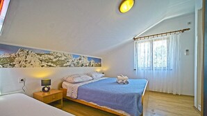 Ein Schlafzimmer mit zwei Betten, einem Gemälde einer Berglandschaft, einem Nachttisch mit Lampe und einem Fenster mit Vorhängen.