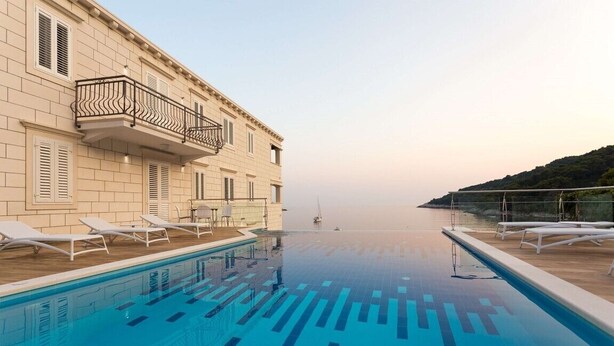 A poolside area with lounge chairs, a clear view of the sea, and a building with balconies.