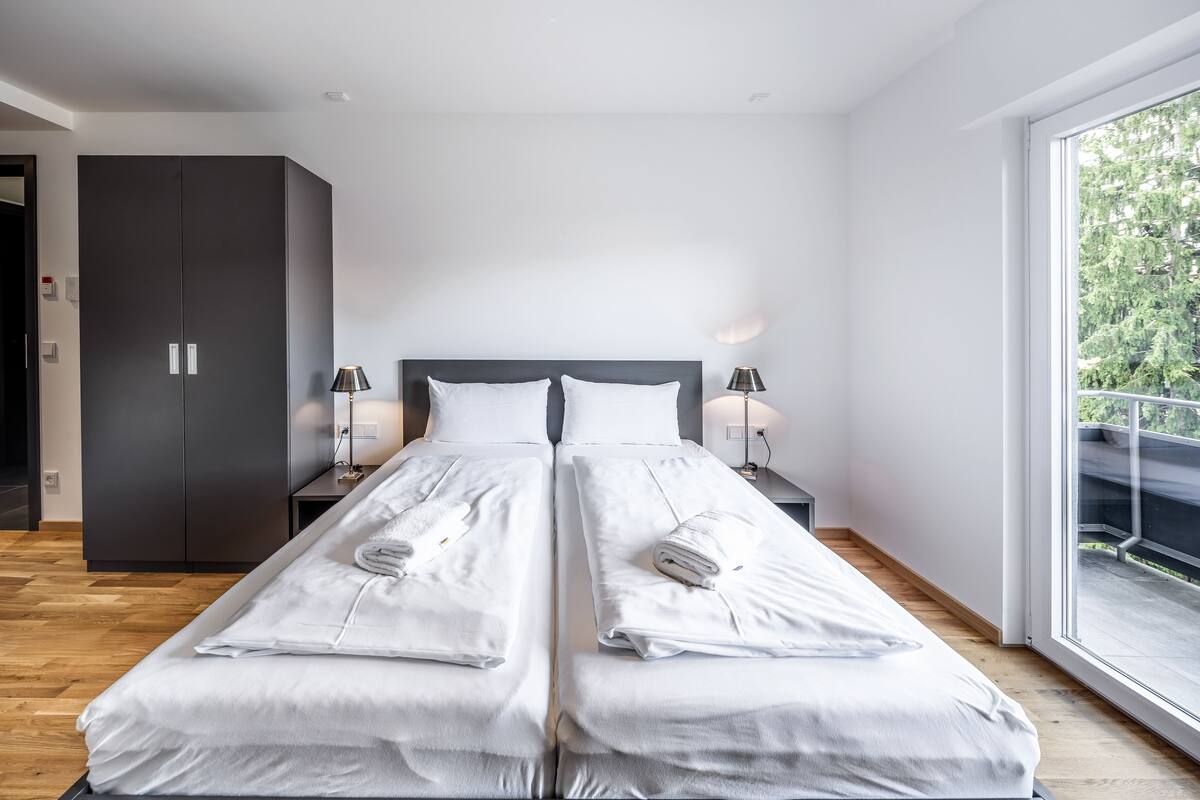 A modern bedroom with a large bed, two bedside tables, a wardrobe, and a balcony with a railing.