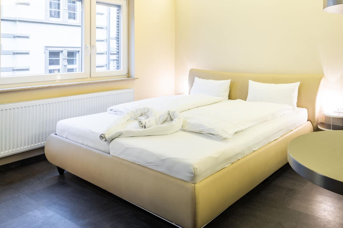 A hotel room with a bed, a bedside table, a window, and a radiator.