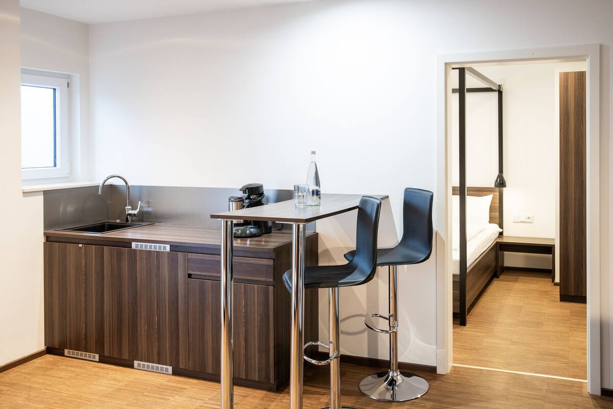 A compact kitchen with a sink, a small island, and two bar stools. There is a window above the sink and a door leading to a bedroom.