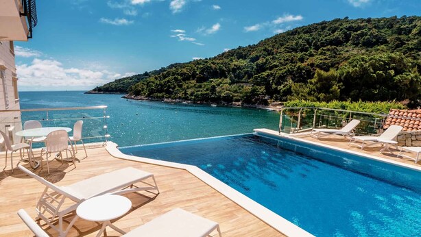 Zona junto a la piscina con sillones reclinables y una mesa, con vistas a una piscina azul cristalina y una ladera boscosa.