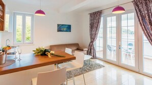 Una cocina moderna con encimera de madera, sillas blancas y una mesa de comedor. Hay dos lámparas colgantes, un ventanal con vistas y un sofá en la zona de estar contigua.