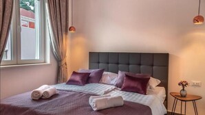 Een net opgemaakt bed met een donker hoofdeinde, twee opgerolde handdoeken en een tafeltje met een bloemenvaas.