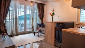 Een moderne woonkamer met een bank, een salontafel en een eethoek met een houten tafel en barkrukken. Er is een groot raam met gordijnen, een vaas met bloemen en een hanglamp.