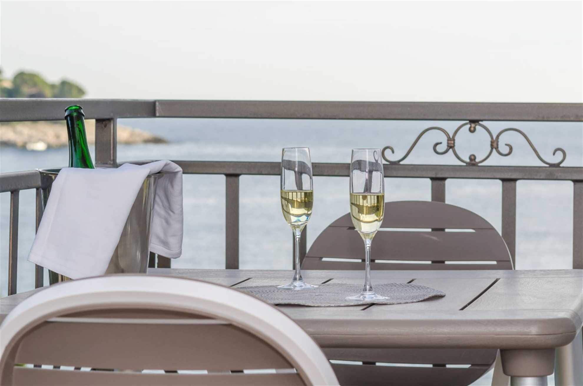 A table with two glasses of white wine, a green bottle, and a white cloth on a balcony overlooking a body of water.