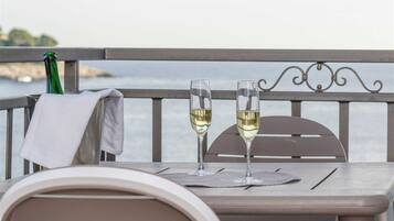 A table with two glasses of white wine, a green bottle, and a white cloth on a balcony overlooking a body of water.