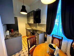 A compact kitchen with a sink, stove, and cabinets, a dining table with chairs, and a window with curtains.