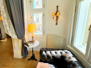 A room with a leather sofa, a table with a lamp, a window with curtains, and a wall-mounted light fixture.