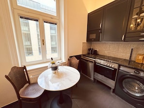 A compact kitchen with a dining table, chairs, a washing machine, and a window with blinds.