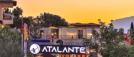 Un hotel llamado "ATALANTE" con fachada de piedra, un balcón y un letrero con un símbolo de sombrero de copa.
