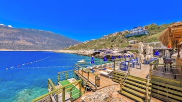 A coastal hotel with outdoor seating, a wooden deck, and a view of the sea and mountains.