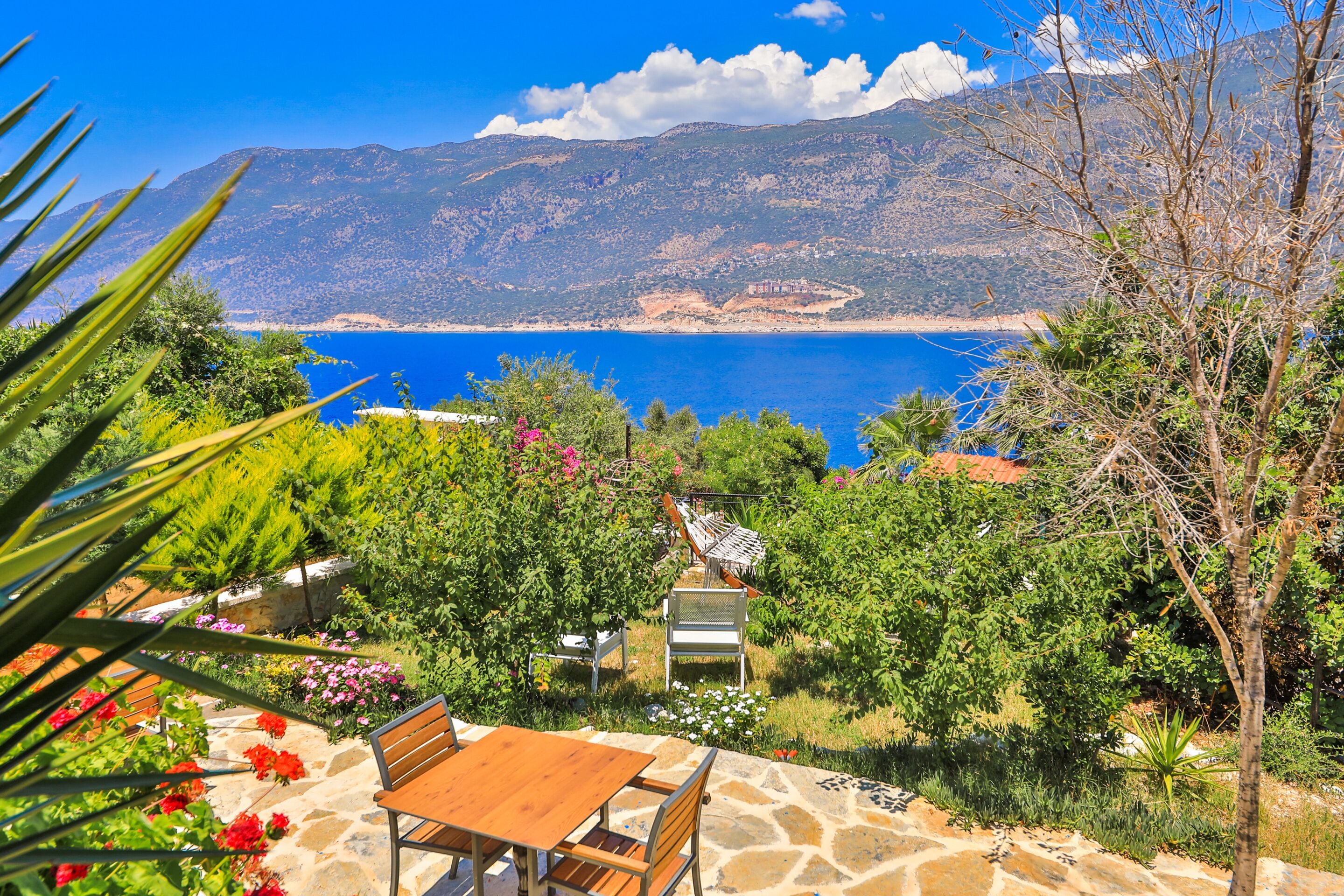 A patio with a table and chairs, overlooking a coastal landscape with mountains and a clear blue sea.