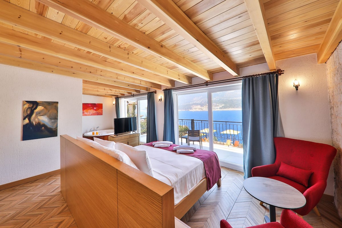 A hotel room with a wooden ceiling, a bed with white linens, a red armchair, a small table, and a balcony with a view of the sea.