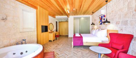 A modern room with a wooden ceiling, a large bathtub, a bed with white bedding, a red armchair, and a small table.