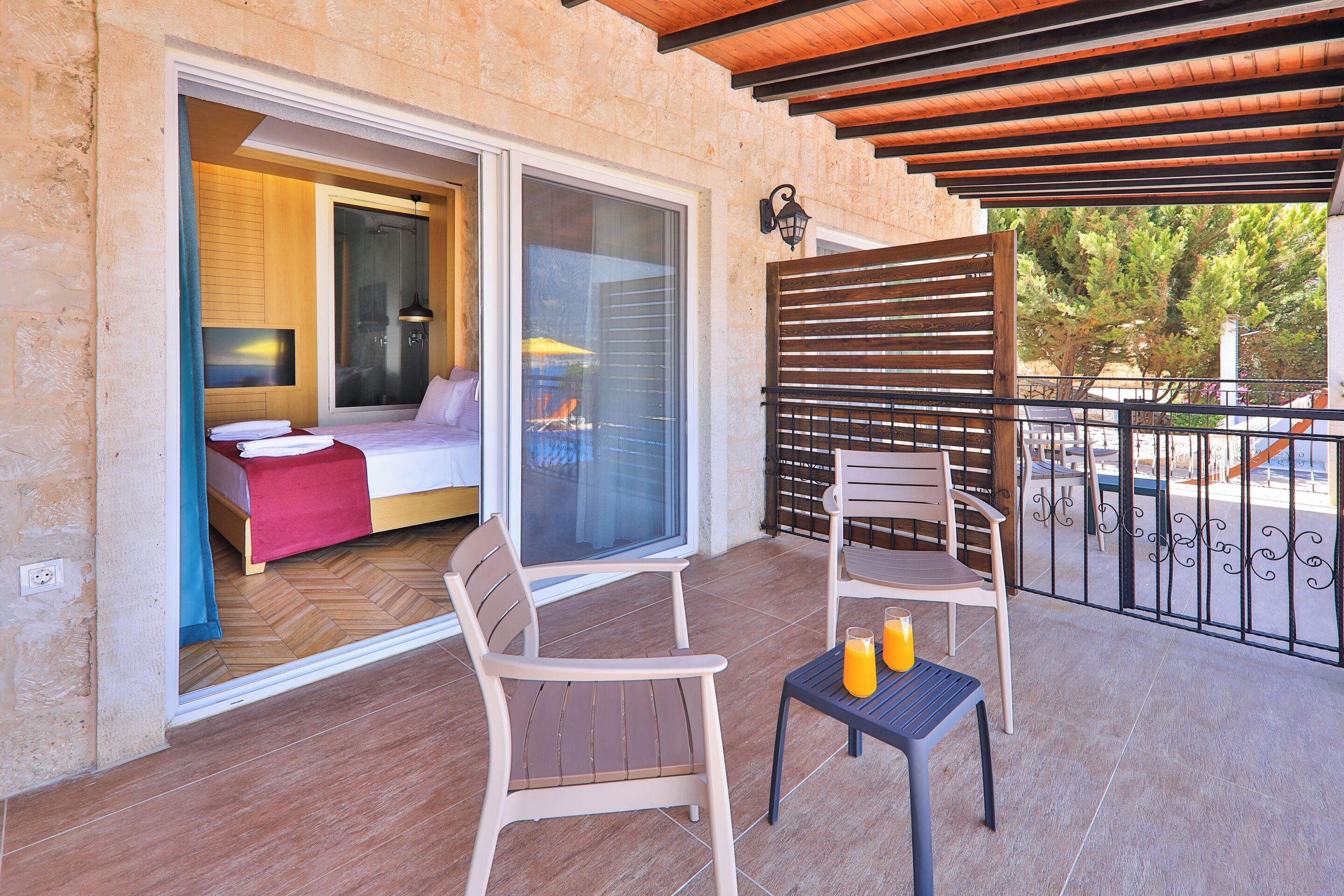 A balcony with two chairs and a table, a sliding glass door leading to a bedroom, and a wooden pergola overhead.