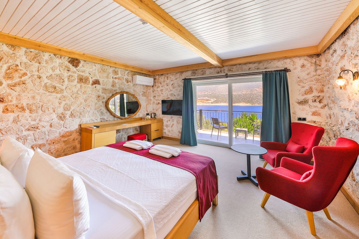 A bedroom with a stone wall, a wooden ceiling, a bed with white linens, a red armchair, a small table, and a TV.