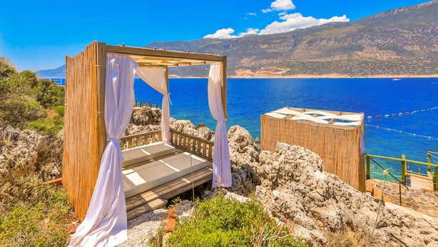 A wooden gazebo with a bed, overlooking a coastal area with a clear blue sea and distant mountains.