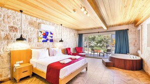 A modern hotel room with a wooden ceiling, stone walls, a large bed, a red chair, a small table, and a hot tub.