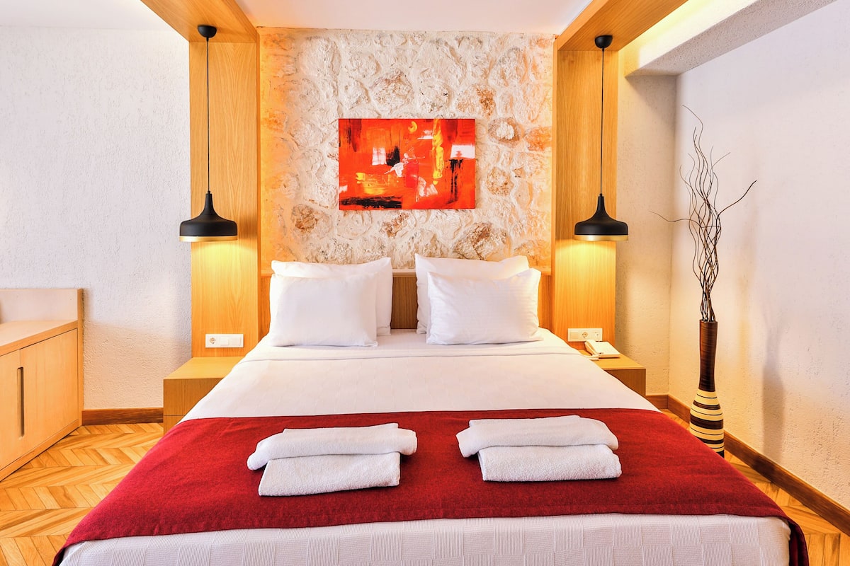 A hotel room with a bed, two bedside tables, a red throw blanket, white pillows, a wall-mounted artwork, and a vase with branches.