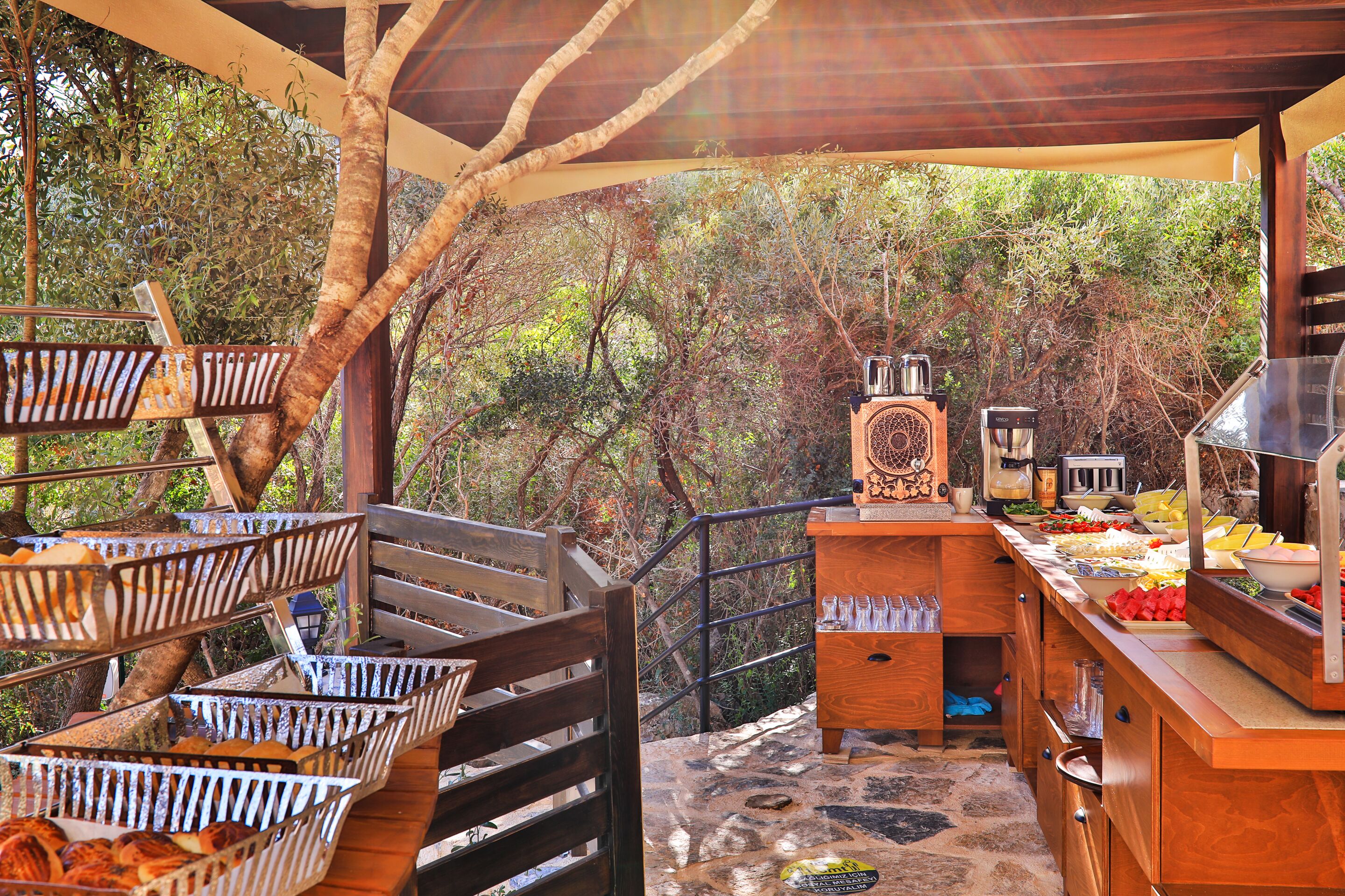 A buffet setup with various food items and a coffee station, set against a backdrop of trees and a wooden deck.