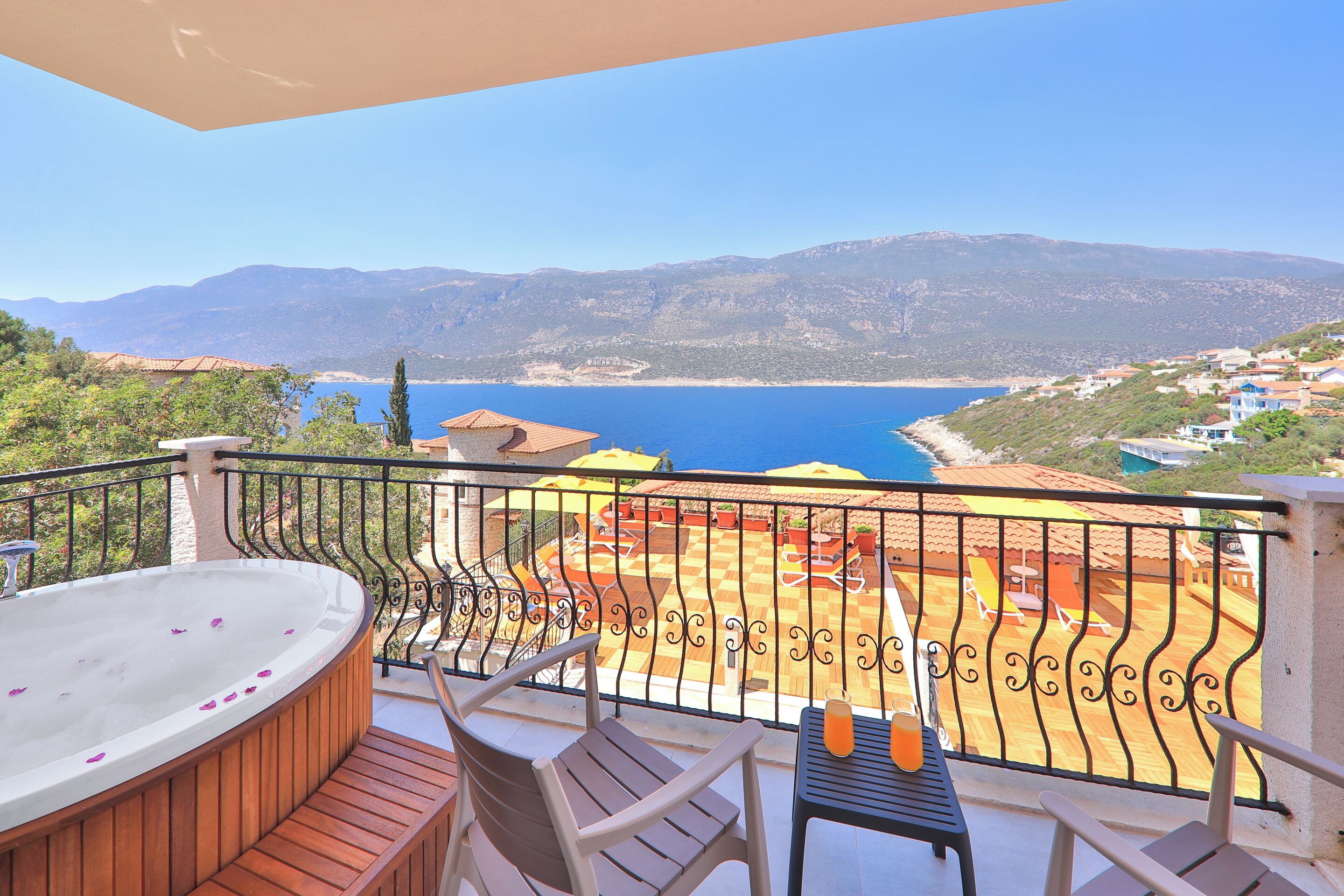 Un balcón con un jacuzzi, sillas de madera y una mesa con una bebida. Disfruta de vistas al área de la piscina con sombrillas y al mar y las montañas.