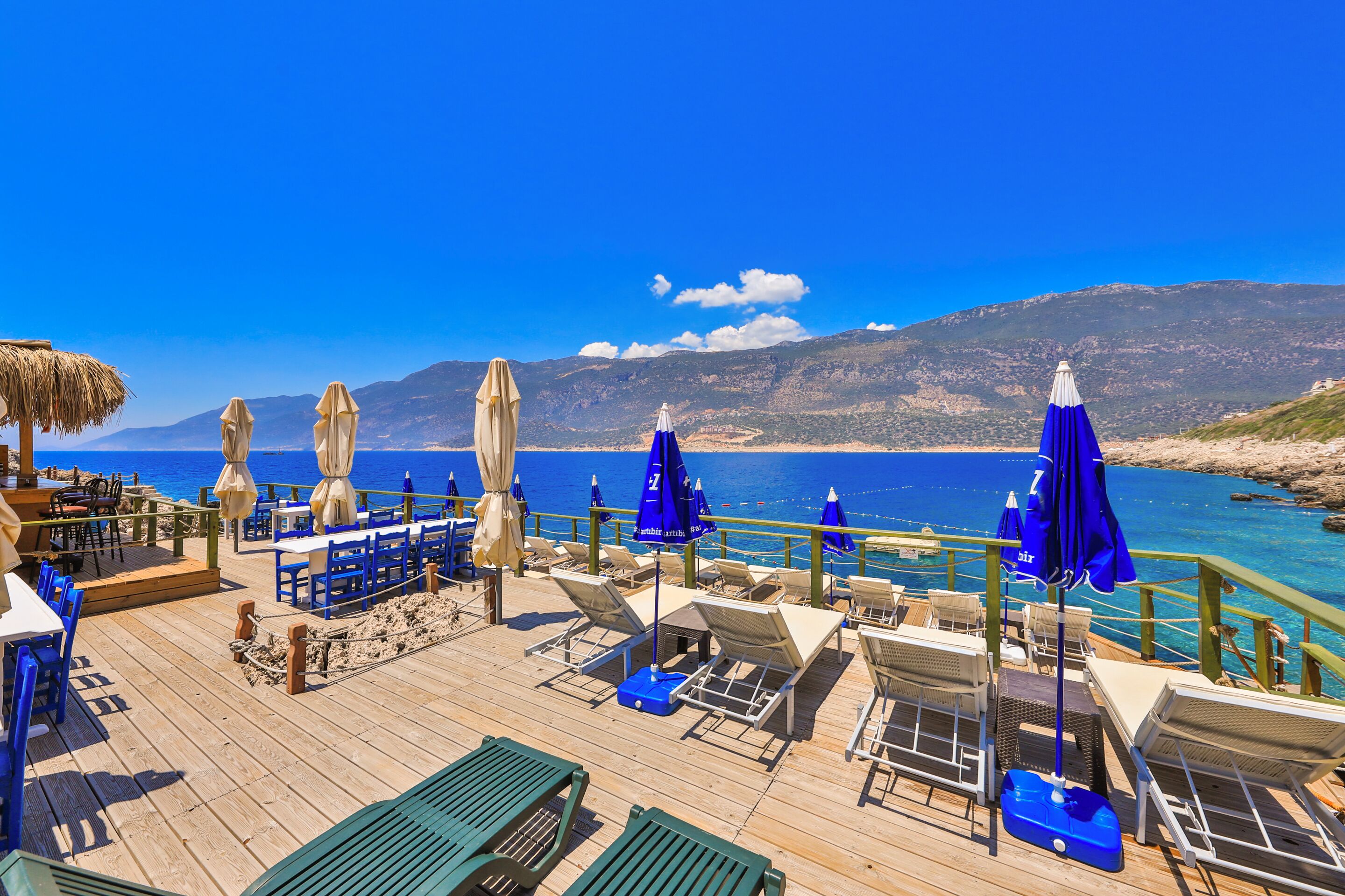 A wooden deck with lounge chairs and umbrellas overlooking a beach and mountains.