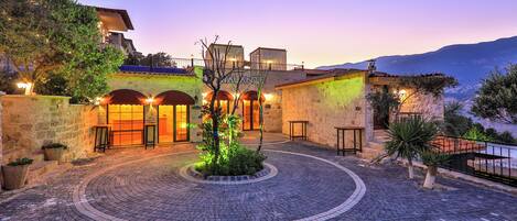 Un patio con un restaurante llamado "Ria Quintana", que cuenta con una pared de piedra, un diseño circular de ladrillos y un pequeño árbol en el centro.