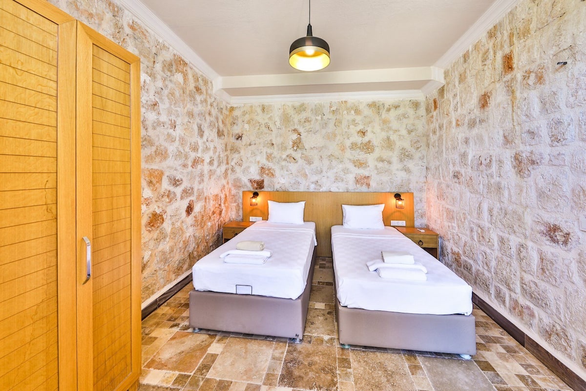 A room with two beds, a wooden door, stone walls, and a ceiling light.
