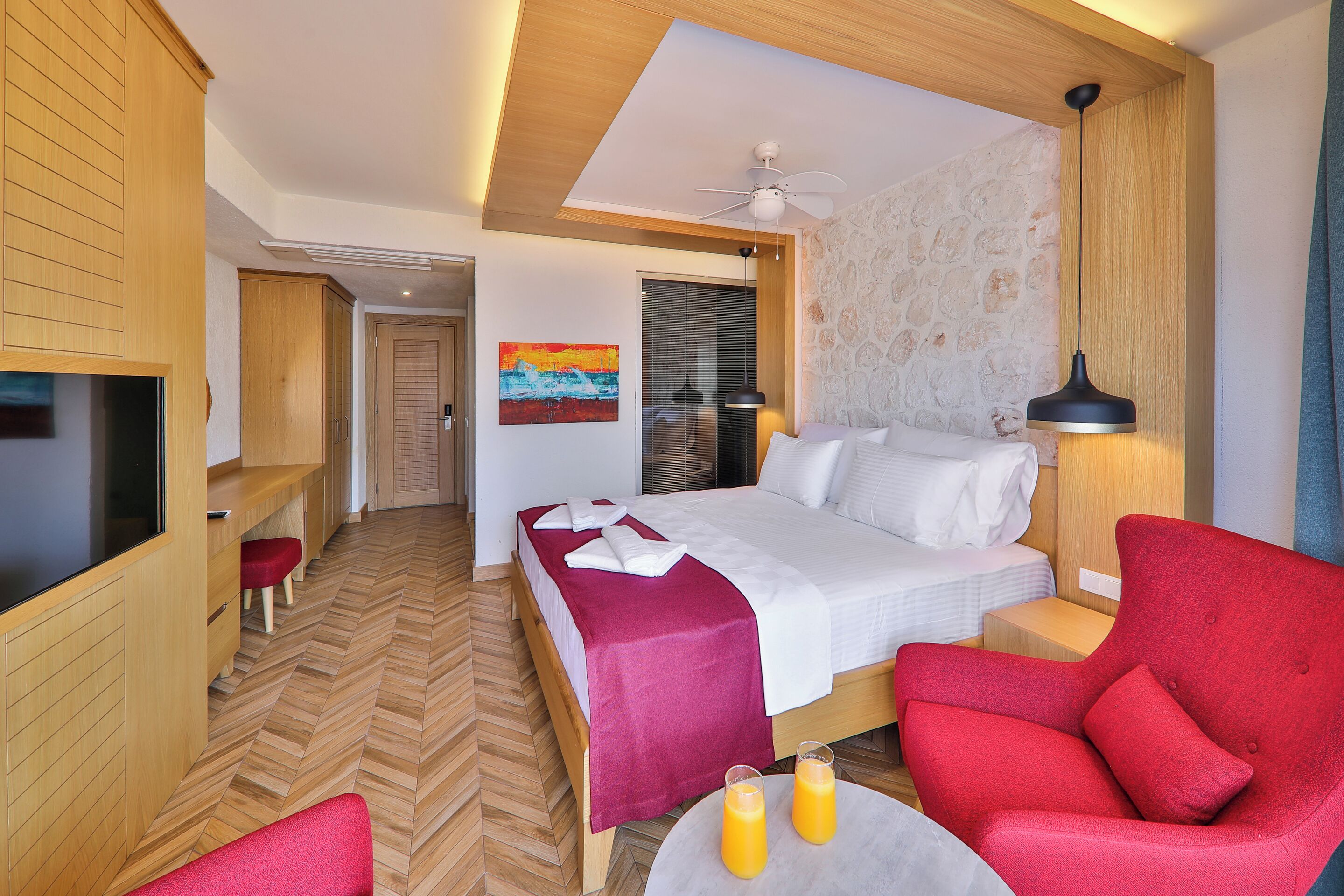 A hotel room with a bed, a red armchair, a small table with drinks, a TV, and a painting on the wall.