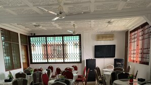 Uma sala de conferências com várias mesas e cadeiras, um ventilador de teto e uma janela grande com grades.