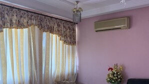 Uma sala de estar com um ventilador de teto, cortinas florais, um sofá bege, um sofá azul, um sofá escuro e uma pequena mesa com um vaso de flores.