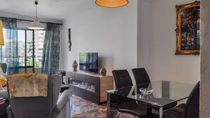 Un salon moderne avec une table à manger en verre, des canapés gris, un téléviseur et un grand tableau encadré au mur.