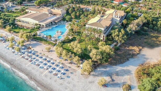 An aerial view of a beachfront property with a swimming pool, beach umbrellas, and a large building with a flat roof.