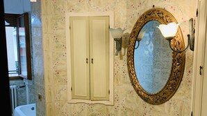 Un baño con baldosas de motivos florales, un lavamanos blanco, un espejo redondo con un marco decorativo y una araña.