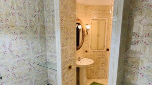 Un baño con paredes de estampado floral, un lavabo blanco, un espejo con marco dorado y una alfombra verde.