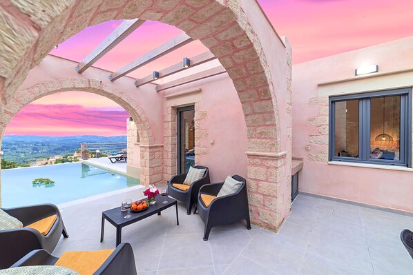 A terrace with a stone archway, two black chairs with cushions, a small table with a vase and oranges, and a view of the landscape.
