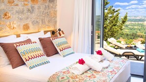 Habitació d'hotel amb paret de pedra, llit amb coixins estampats, vista del paisatge a través de grans finestres i un balcó amb gandules.
