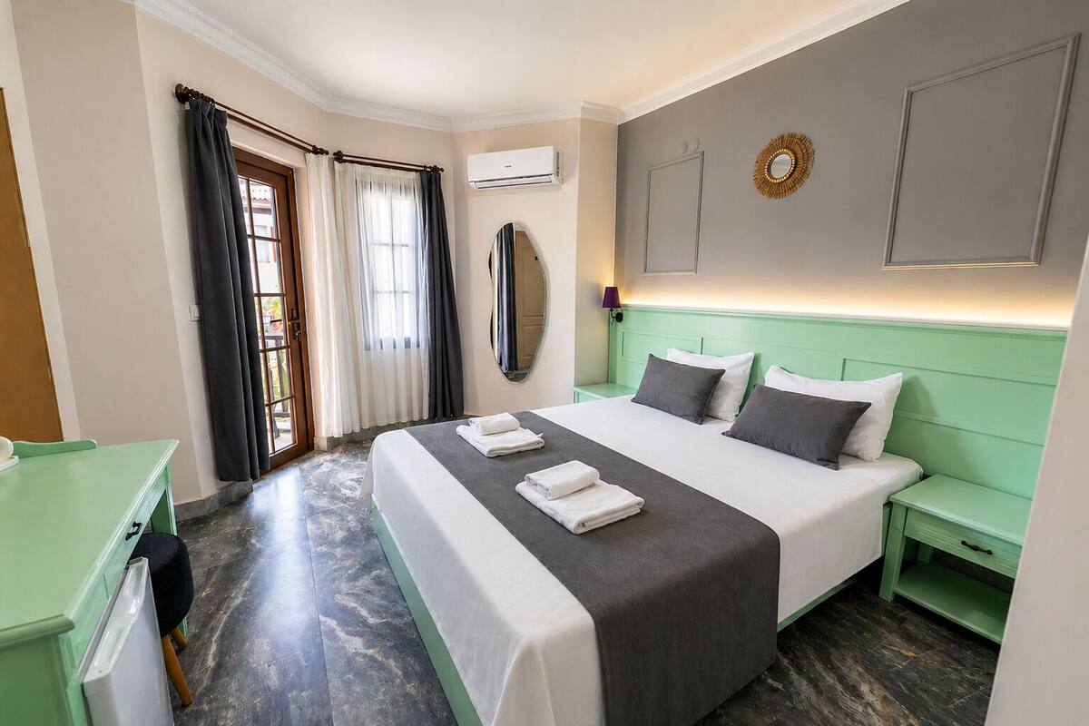 A hotel room with a bed, a desk, a mirror, a wall-mounted air conditioner, and a window with curtains.