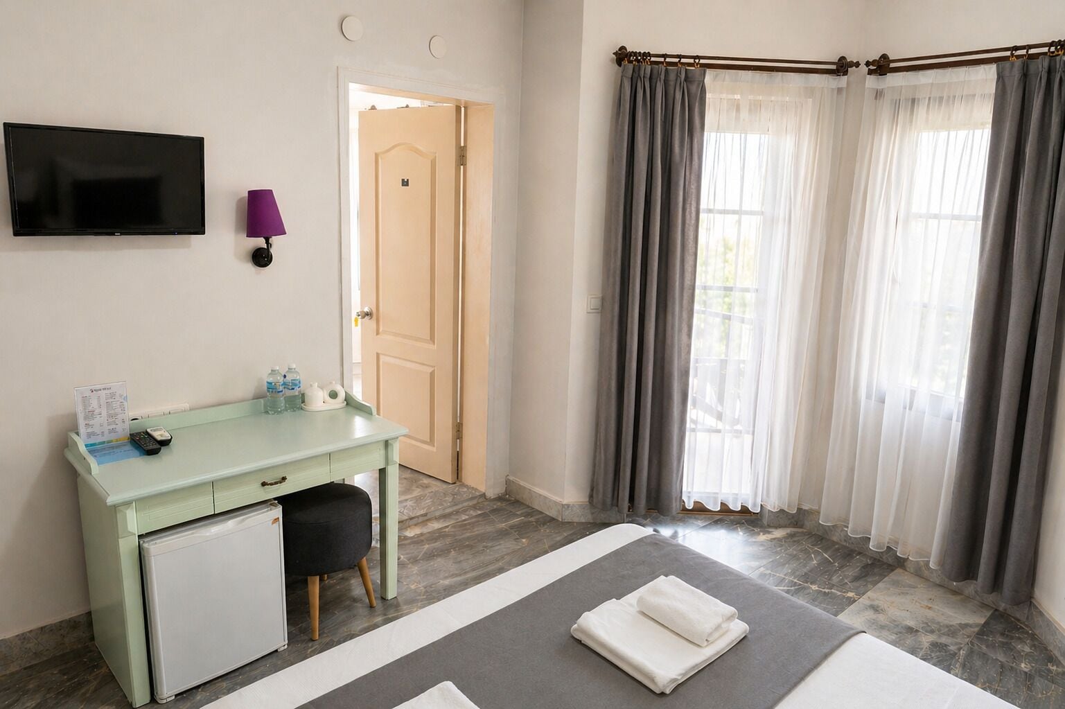 Habitación de hotel con escritorio, televisión, puerta y ventana con cortinas.